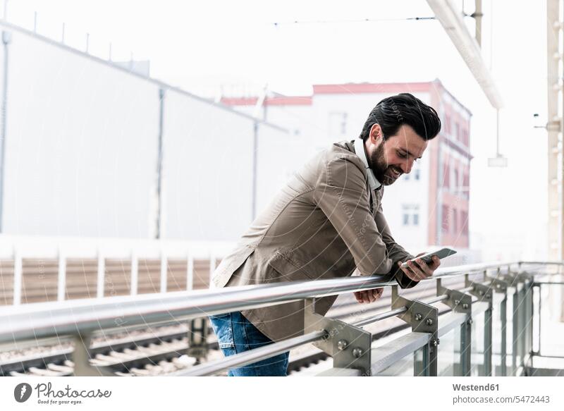 Lächelnder junger Mann mit Handy auf dem Bahnsteig Bahnhof Bahnhöfe Bahnhoefe Zugbahnsteig Zugbahnsteige Bahnsteige lächeln Mobiltelefon Handies Handys