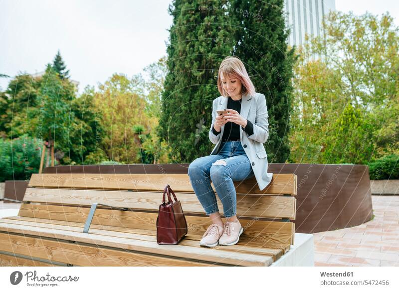 Lächelnde Frau sitzt auf Parkbank und benutzt Mobiltelefon geschäftlich Geschäftsleben Geschäftswelt Geschäftsperson Geschäftspersonen Businessfrau