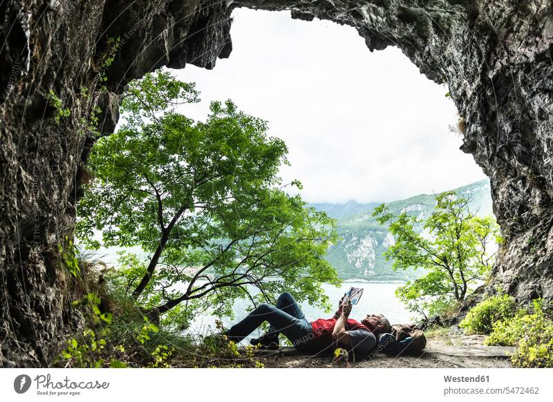 Ein erwachsener Mann liest ein Buch, während er am Höhleneingang am Comer See liegt Farbaufnahme Farbe Farbfoto Farbphoto Comersee Comosee Lario Lago di Como