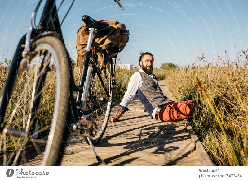 Gut gekleideter Mann sitzt auf einem Holzsteg in der Landschaft neben einem Fahrrad Leute Menschen People Person Personen Europäisch Kaukasier kaukasisch 1 Ein