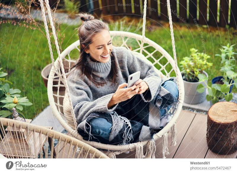 Glückliche junge Frau benutzt ein Smartphone, während sie im Garten auf der Schaukel sitzt Europäer Europäisch Kaukasier kaukasisch eine Person ein Mensch
