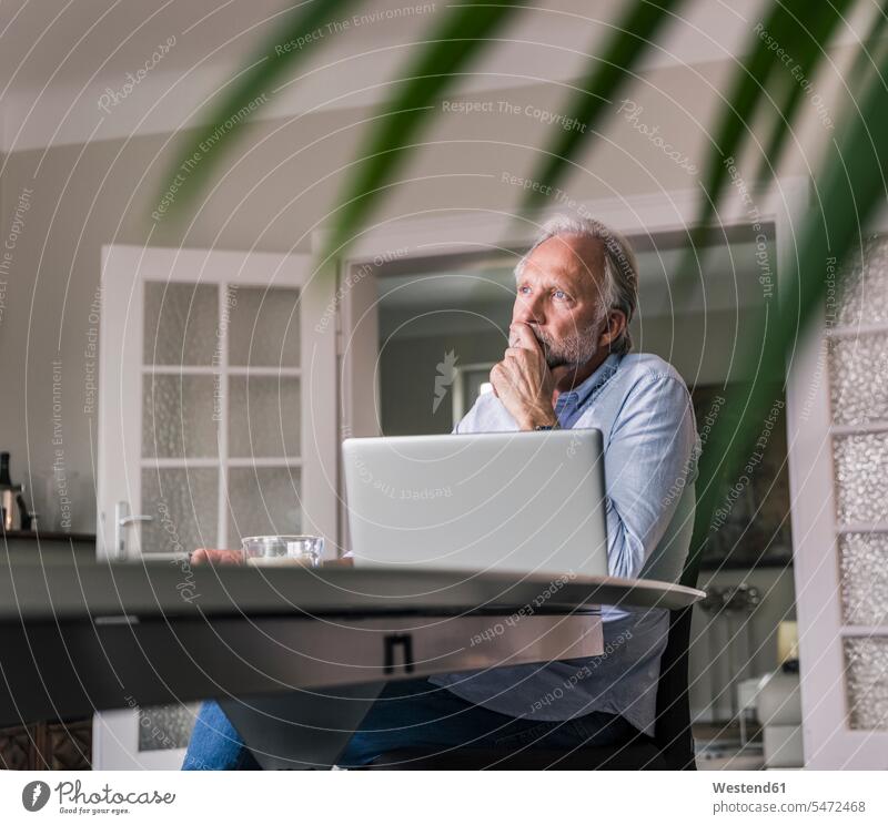Nachdenklicher reifer Mann sitzt am Tisch mit Laptop in seinem Wohnzimmer nachdenklich Notebook Laptops Notebooks Männer männlich Tische sitzen sitzend Wohnraum