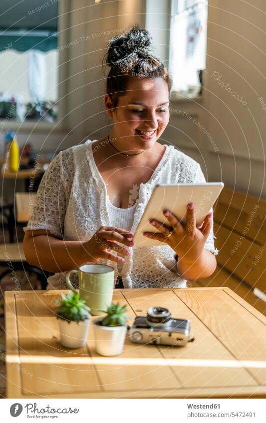 Lächelnde junge Frau benutzt digitales Tablet, während sie in einem Café am Tisch sitzt Farbaufnahme Farbe Farbfoto Farbphoto Innenaufnahme Innenaufnahmen innen