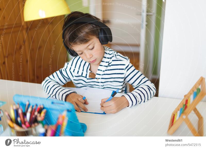 Junge macht Hausunterricht und schreibt am Notebook, benutzt Tablett und Kopfhörer zu Hause Tische Arbeitstisch Schreibtische Stifte Kopfhoerer hoeren
