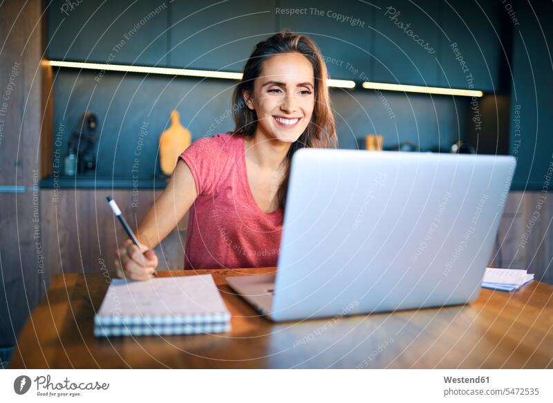 Lächelnde Unternehmerin schaut auf den Laptop, während sie im Notizblock auf dem Schreibtisch schreibt Farbaufnahme Farbe Farbfoto Farbphoto Freizeitkleidung