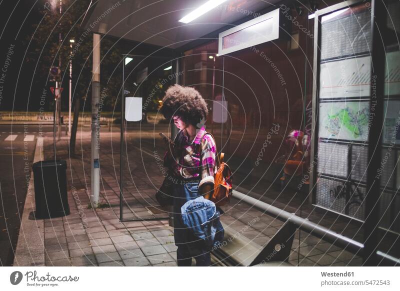 Junge Frau mit Afro-Frisur benutzt Smartphone an Bushaltestelle in der Stadt Leute Menschen People Person Personen gelockt gelockte Haare gelocktes Haar lockig