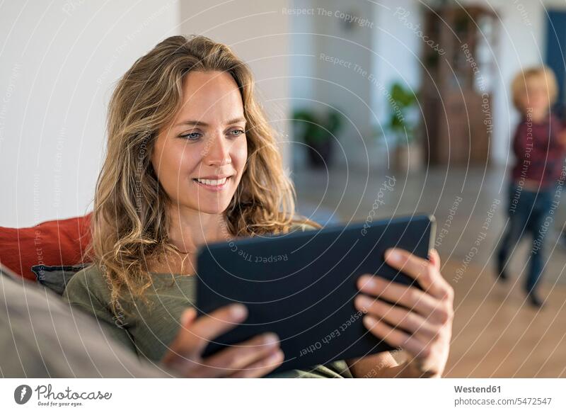 Lächelnde Frau realxing auf Couch zu Hause mit Tablet mit Sohn im Hintergrund Tablet Computer Tablet-PC Tablet PC iPad Tablet-Computer weiblich Frauen Zuhause