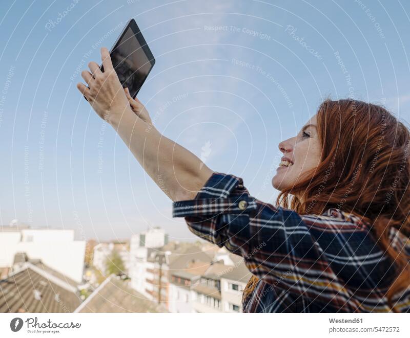 Lächelnde rothaarige Frau mit Tablet auf der Dachterrasse, die ein Selfie macht Leute Menschen People Person Personen Europäisch Kaukasier kaukasisch 1 Ein