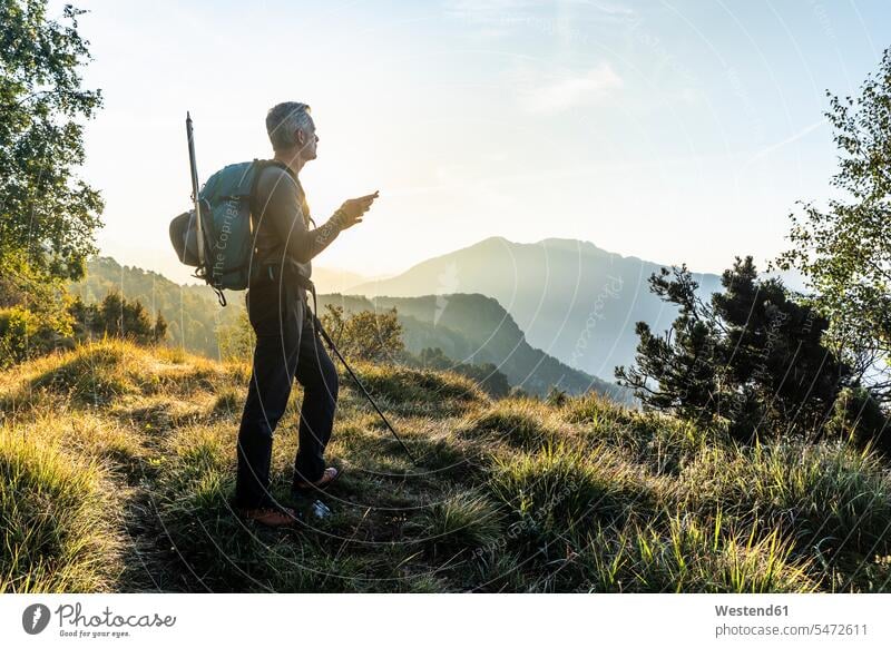 Mann benutzt Mobiltelefon, während er bei Sonnenaufgang auf einem Berg gegen den Himmel steht, Orobie, Lecco, Italien Farbaufnahme Farbe Farbfoto Farbphoto