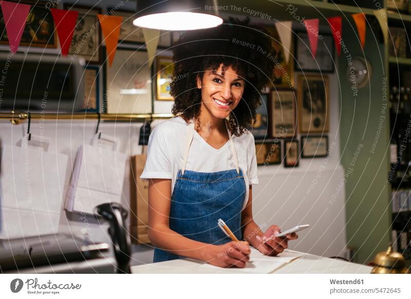 Porträt einer lächelnden Frau mit Handy und Notizbuch in einem Geschäft Notizbücher Notizbuecher Shop Laden Läden Geschäfte Shops Mobiltelefon Handies Handys