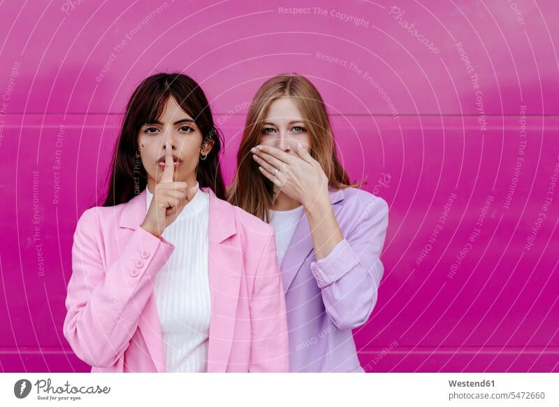 Junge Frau mit Finger auf den Lippen steht neben Schwester und bedeckt Mund gegen rosa Wand Farbaufnahme Farbe Farbfoto Farbphoto Außenaufnahme außen draußen