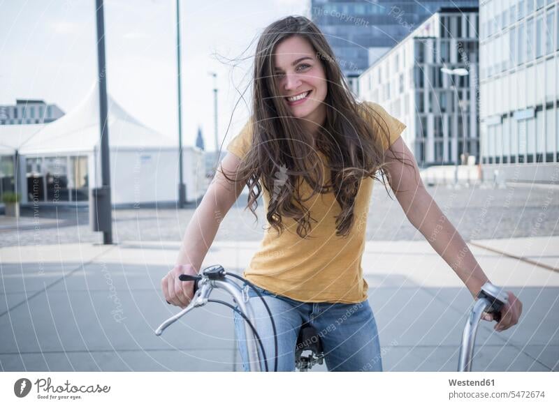 Deutschland, Köln, Porträt einer lächelnden jungen Frau mit wehenden Haaren auf ihrem Fahrrad Europäer Kaukasier kaukasisch Europäisch Außenaufnahme außen