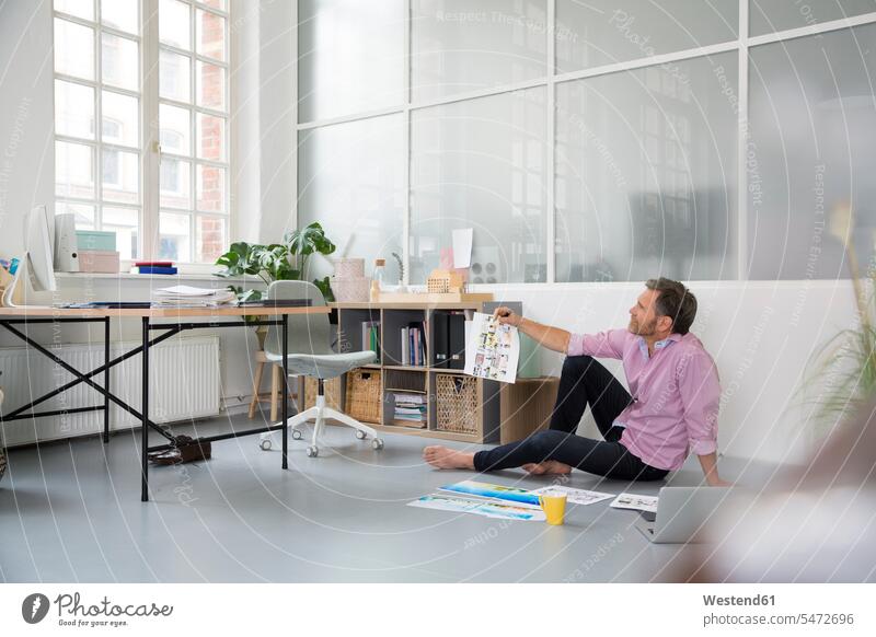 Lässiger Mann mit Plänen und Laptop, der in einem Loft-Büro auf dem Boden sitzt Office Büros Notebook Laptops Notebooks Männer männlich leger sitzen sitzend