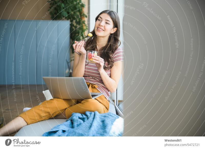 Junge Frau sitzt am Fenster zu Hause mit Laptop und isst einen Snack lächeln Imbiss Zwischenmahlzeit Snacks weiblich Frauen Zuhause daheim sitzen sitzend essen