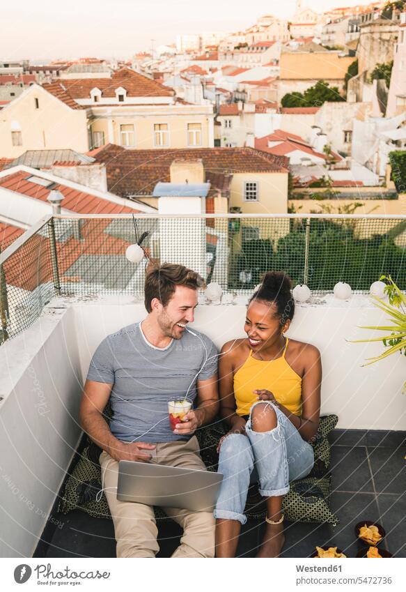 Glückliches junges Paar mit Laptop und Kopfhörern sitzt abends auf dem Dach, Lissabon, Portugal Leute Menschen People Person Personen Afrikanisch
