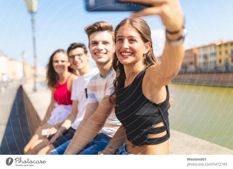 Italien, Pisa, Gruppe von vier glücklichen Freunden, die auf einer Mauer am Arno sitzen und ein Selfie machen sitzend sitzt Mauern Selfies Gemeinsam Zusammen
