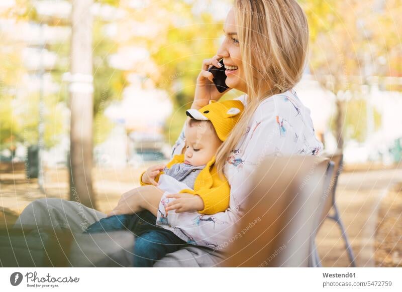 Glückliche Mutter ruht sich mit dem kleinen Jungen auf einer Parkbank aus und telefoniert Jacken Bänke Sitzbank Sitzbänke Parkbänke Telekommunikation