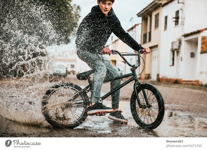 Zuversichtlicher Teenager, der in der Regenzeit beim Radfahren auf der Straße Wasser in einer Pfütze verspritzt Farbaufnahme Farbe Farbfoto Farbphoto