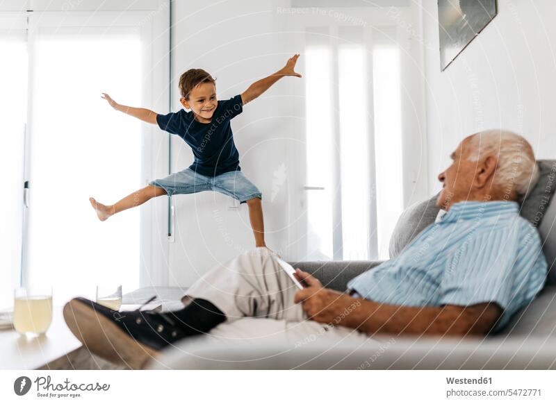 Großvater sitzt mit Tablet auf dem Sofa und beobachtet seinen Enkel, der in die Luft springt zuschauen beobachten anschauen betrachten Enkelsöhne Enkelsohn Opa