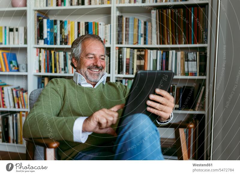 Lächelnder Mann benutzt digitales Tablett, während er zu Hause am Bücherregal sitzt Farbaufnahme Farbe Farbfoto Farbphoto Innenaufnahme Innenaufnahmen innen