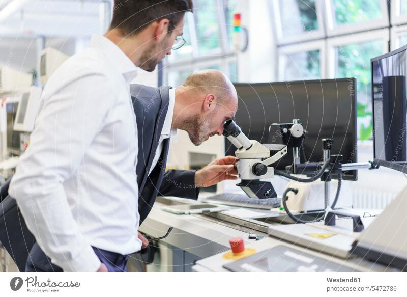Junger männlicher Techniker, der einem Geschäftsmann zur Seite steht und im Labor durch ein Mikroskop schaut Farbaufnahme Farbe Farbfoto Farbphoto Innenaufnahme