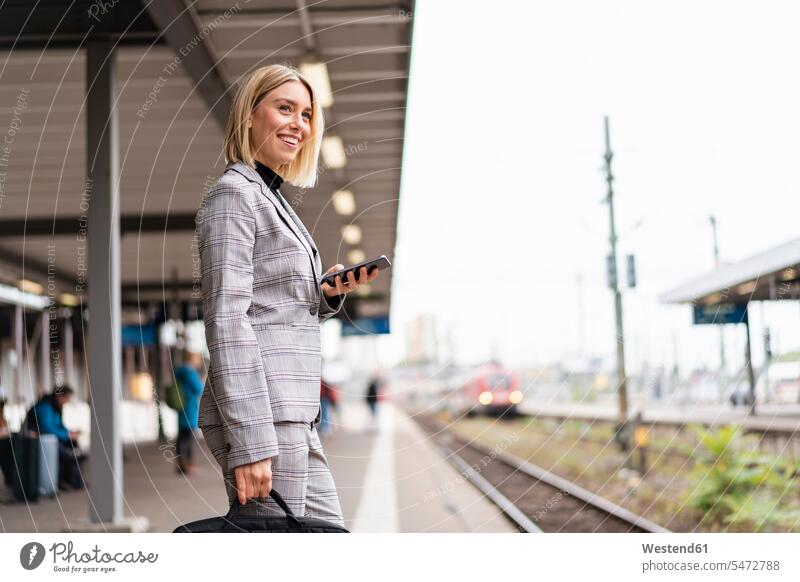 Lächelnde junge Geschäftsfrau mit Mobiltelefon am Bahnhof geschäftlich Geschäftsleben Geschäftswelt Geschäftsperson Geschäftspersonen Businessfrau