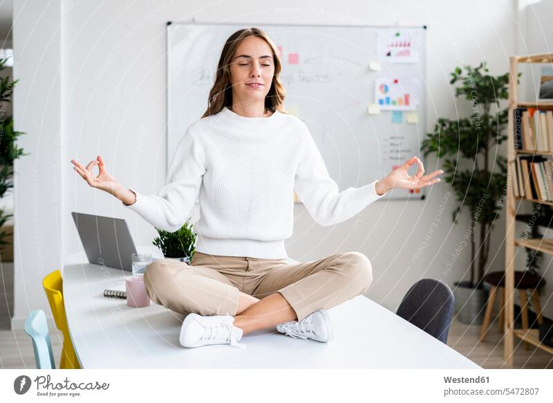 Junge Geschäftsfrau praktiziert Yoga, während sie im Büro am Tisch sitzt Farbaufnahme Farbe Farbfoto Farbphoto Innenaufnahme Innenaufnahmen innen drinnen Tag
