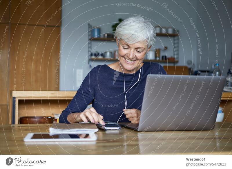Lächelnde Frau hört Musik auf einem Smartphone und benutzt einen Laptop, während sie zu Hause sitzt Farbaufnahme Farbe Farbfoto Farbphoto Innenaufnahme
