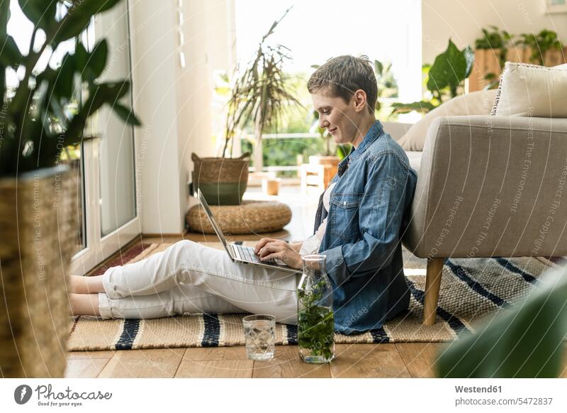 Mittlere erwachsene Frau mit kurzen Haaren, die einen Laptop benutzt, während sie zu Hause auf einem Teppich sitzt Farbaufnahme Farbe Farbfoto Farbphoto