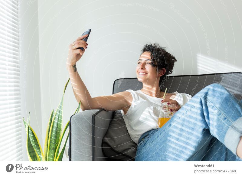 Lächelnde junge Frau sitzt mit einem Softdrink auf der Couch und macht ein Selfie Leute Menschen People Person Personen gelockt gelockte Haare gelocktes Haar