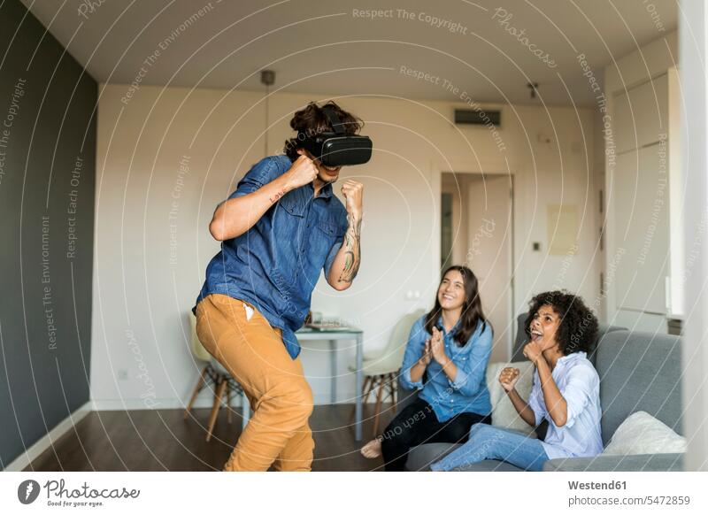 Zwei Frauen ermutigen Mann beim Spielen mit VR-Brille zu Hause Zuhause daheim Freunde anfeuern spielen Virtuelle Realität Virtuelle Realitaet Brillen
