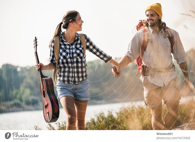 Glückliches junges Paar mit Rucksäcken und Gitarre beim Spaziergang am Flussufer Rucksack glücklich glücklich sein glücklichsein Fluesse Fluß Flüsse Pärchen