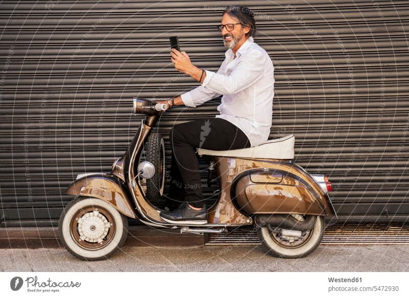 Glücklicher reifer Mann, der sich auf einem Motorroller gegen einen schwarzen Fensterladen setzt Farbaufnahme Farbe Farbfoto Farbphoto Außenaufnahme außen