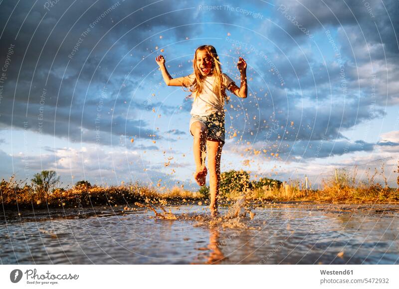 Fröhliches Mädchen spritzt Wasser auf Pfütze gegen bewölkten Monsunhimmel Farbaufnahme Farbe Farbfoto Farbphoto Spanien Außenaufnahme außen draußen im Freien