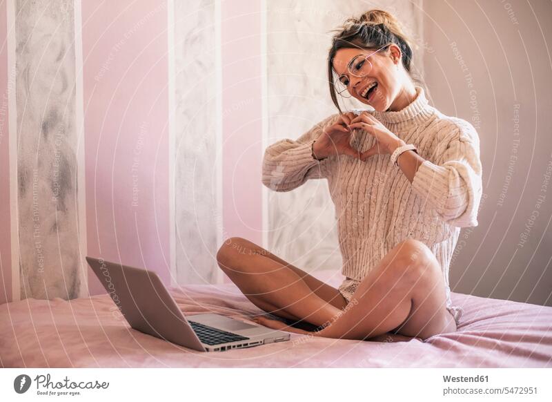 Junge Frau sitzt auf dem Bett mit Laptop bilden ein Herz mit ihren Händen während Video-Chat Betten Notebook Laptops Notebooks Herzform Herzformen Herzen