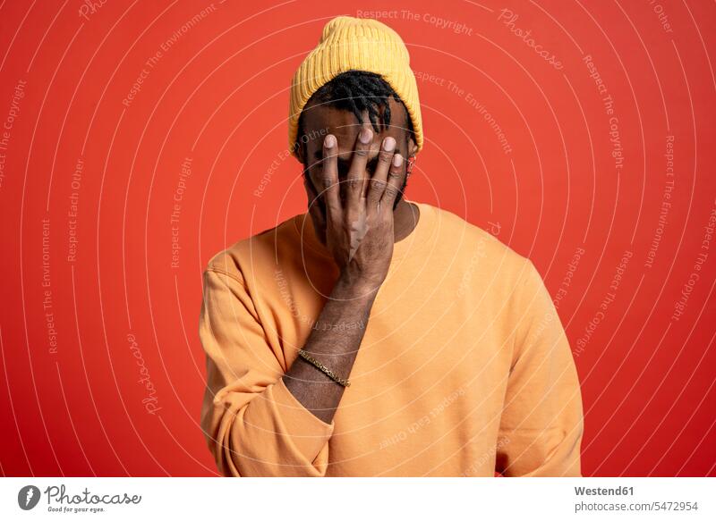 Junger Mann bedeckt sein Gesicht vor orangefarbener Wand pessimistisch schwarzseherisch gefühlvoll Emotionen Empfindung Empfindungen fühlen Gefühl Gefühle