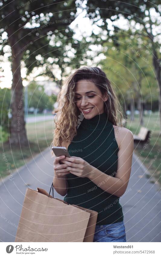 Lächelnde Frau mit Smartphone im öffentlichen Park Farbaufnahme Farbe Farbfoto Farbphoto Außenaufnahme außen draußen im Freien Tag Tageslichtaufnahme