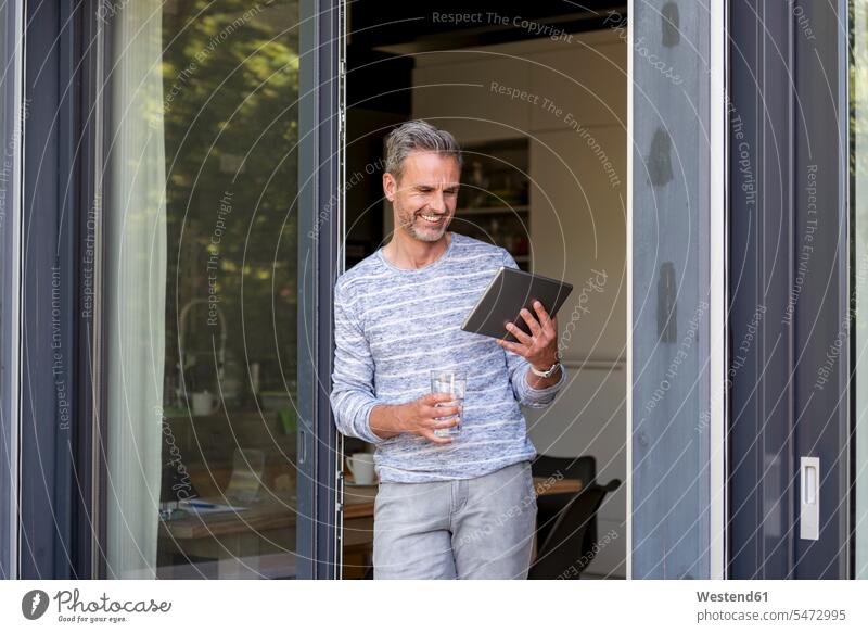 Lächelndes reifes Stehen an der französischen Tür zu Hause mit einer Tablette stehen stehend steht Fenstertür Zuhause daheim lächeln Mann Männer männlich