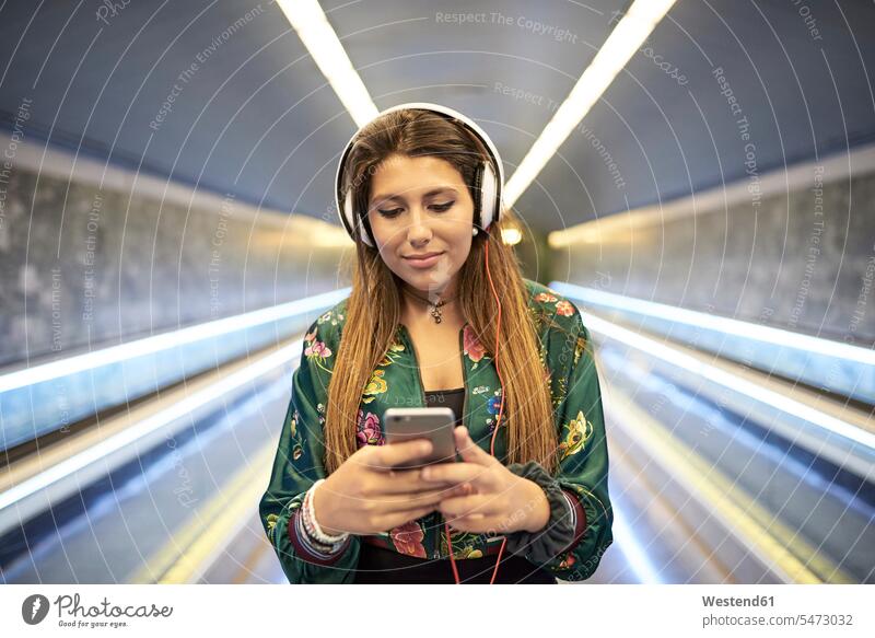 Porträt einer Frau mit Kopfhörern, die in der U-Bahn-Station auf ihr Handy schaut U-Bahnstation U-Bahnstationen U-Bahnhöfe U-Bahnhoefe U-Bahnhof Kopfhoerer