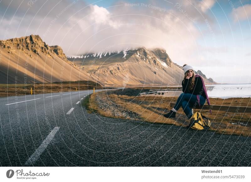 Frau mit Rucksack sitzt auf einem Geländer, während sie am Straßenrand gegen die Berge in Island wartet Farbaufnahme Farbe Farbfoto Farbphoto Außenaufnahme