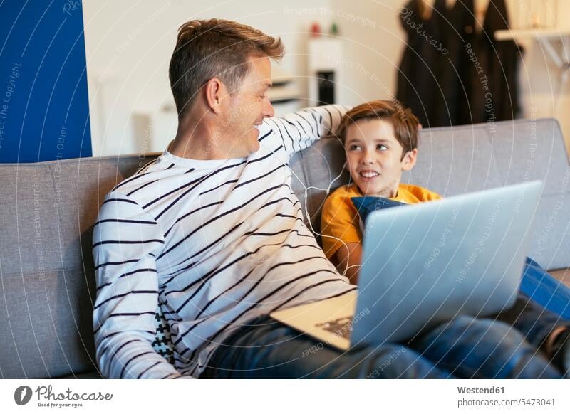 Lächelnder Vater und Sohn mit Ohrstöpseln und Laptop auf der Couch zu Hause Notebook Laptops Notebooks Söhne Sofa Couches Liege Sofas lächeln Papas Väter Vati