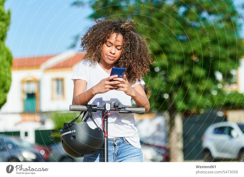 Frau benutzt ein Smartphone, während sie an einem sonnigen Tag mit einem elektrischen Roller steht Farbaufnahme Farbe Farbfoto Farbphoto Außenaufnahme außen