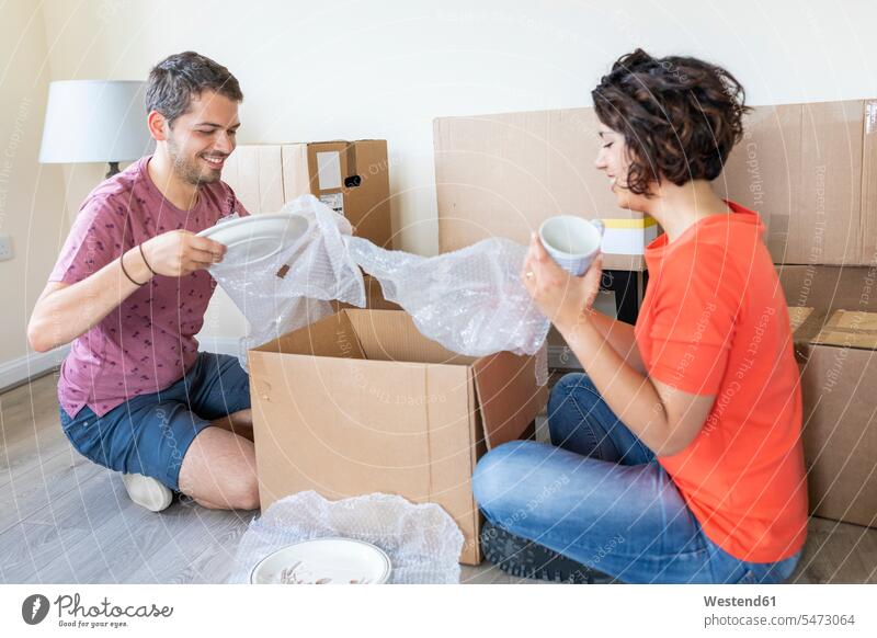 Glückliches Paar zieht in ein neues Zuhause ein und packt einen Karton aus Kartons Pappkarton Pappkartons Folien T-Shirts sitzend sitzt umziehen freuen
