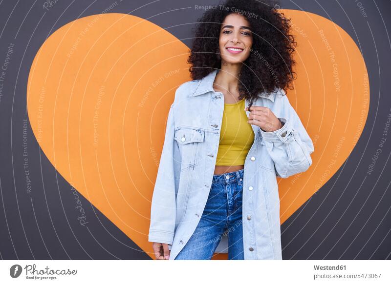 Schöne Frau mit lockigem Haar trägt Jeansjacke stehen gegen Herzform an der Wand Farbaufnahme Farbe Farbfoto Farbphoto Deutschland Freizeitbeschäftigung Muße
