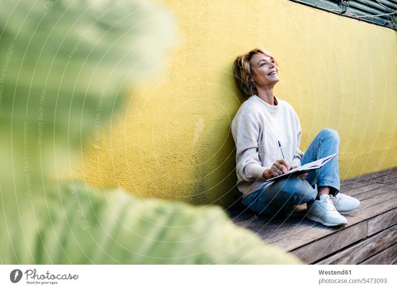Nachdenklich lächelnde Frau sitzt mit Tagebuch gegen gelbe Wand auf Gebäudeterrasse Farbaufnahme Farbe Farbfoto Farbphoto Außenaufnahme außen draußen im Freien