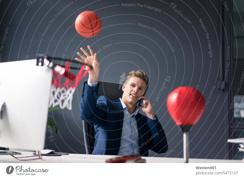 Junger Geschäftsmann sitzt am Schreibtisch im Büro spielen Basketball und sprechen auf Handy Businessmann Businessmänner Geschäftsmänner Office Büros