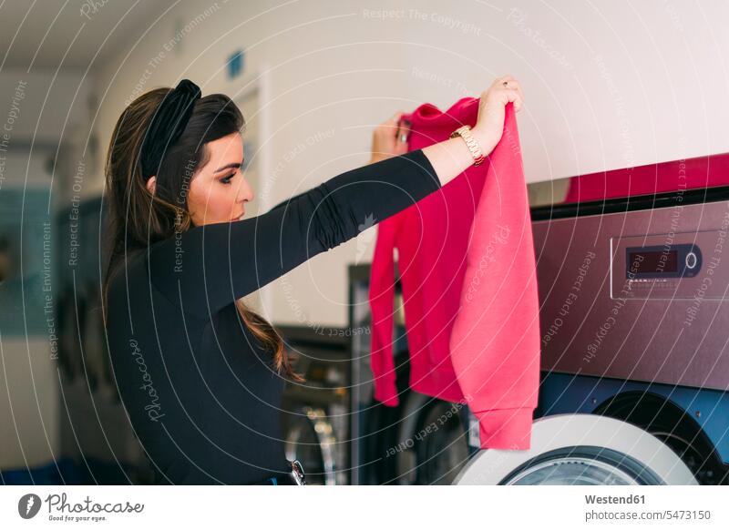 Junge Frau, die in einem Waschsalon die Wäsche wäscht, betrachtet rosa Sweatshirt Leute Menschen People Person Personen Europäisch Kaukasier kaukasisch 1 Ein