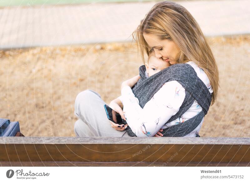 Glückliche Mutter ruht mit dem kleinen Jungen auf einer Parkbank Bänke Sitzbank Sitzbänke Parkbänke knuddeln schmusen sitzend sitzt Arm umlegen Umarmung