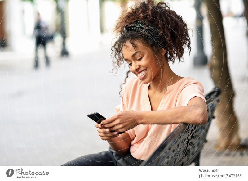 Lächelnde junge Frau sitzt auf einer Bank und benutzt ein Mobiltelefon weiblich Frauen sitzen sitzend glücklich Glück glücklich sein glücklichsein Handy Handies