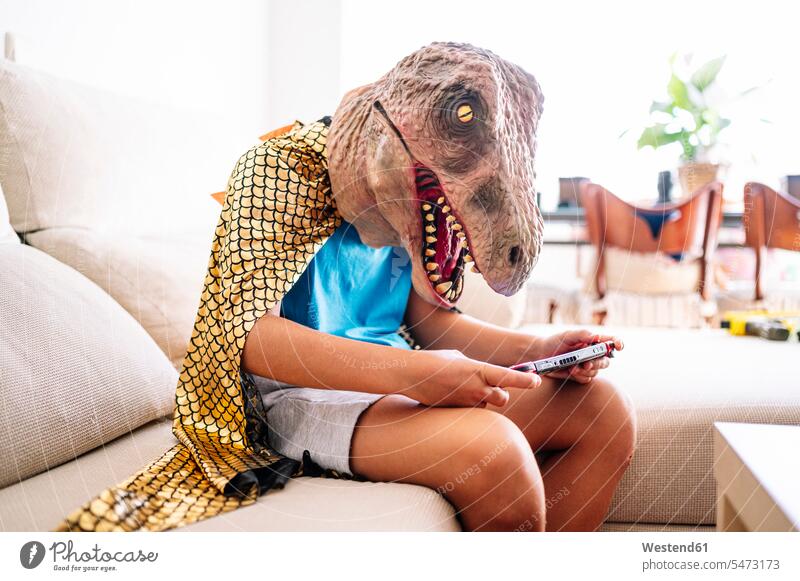 Junge mit Dinosaurier-Maske und Umhang, der ein Smartphone benutzt, während er zu Hause auf dem Sofa sitzt Farbaufnahme Farbe Farbfoto Farbphoto Spanien